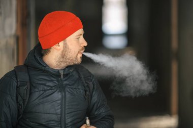 Vape hayatta. Beyaz genç adam kırmızı şapka ve siyah ceket puffs harap bir ev koridorda Elektronik Sigara Buhar kapalı icar.