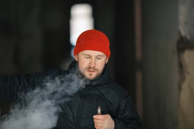 Vape hayatta. Beyaz genç adam kırmızı şapka ve siyah ceket puffs harap bir ev koridorda Elektronik Sigara Buhar kapalı icar.