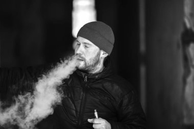 Vape hayatta. Genç beyaz adam bir şapka ve siyah ceket puffs harap bir ev koridorda Elektronik Sigara Buhar kapalı icar. Siyah ve beyaz.