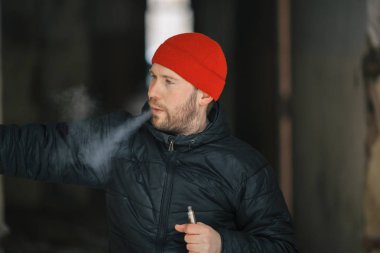Vape hayatta. Beyaz genç adam kırmızı şapka ve siyah ceket puffs harap bir ev koridorda Elektronik Sigara Buhar kapalı icar.