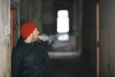 Vape hayatta. Beyaz genç adam kırmızı şapka ve siyah ceket puffs harap bir ev koridorda Elektronik Sigara Buhar kapalı icar.