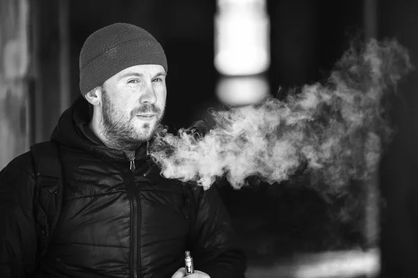 Vape in life. Young white man in a hat and black jacket letting off puffs of steam from electronic cigarette in the corridor of a ruined house. Black and white.