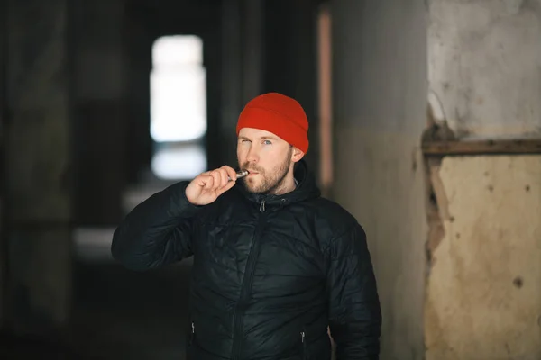 Vape in life. Young white man in red hat and black jacket is smoking an electronic cigarette in the corridor of a ruined house.