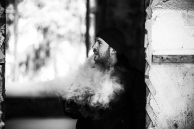 Vape hayatta. Genç beyaz adam kırmızı şapka ve siyah ceket puffs harap bir ev koridorda Elektronik Sigara Buhar kapalı icar. Siyah