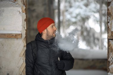 Vape hayatta. Beyaz genç adam kırmızı şapka ve siyah ceket puffs harap bir ev koridorda Elektronik Sigara Buhar kapalı icar.