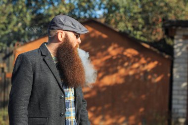 Vape adam gerçek hayatta sakallı. Güneş gözlüğü ve gri kap vaping elektronik sigara sonbahar köyde Caddesi üzerinde büyük sakallı genç adam portresi.
