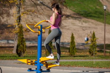 Güneş gözlüklü genç atletik kız sonbaharda Park sokak bir oyun alanı üzerinde bir spor simülatörü yanında duruyor.