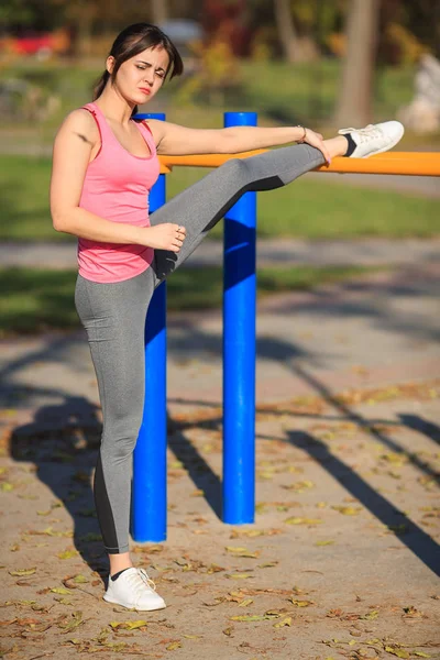 Young athletic girl in a pink t-shirt doing stretching next to gymnastics bars on a street sports ground in the park in the autumn.