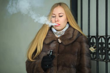 Vape hanım. Bir kürk ceket genç güzel kız sarışın elektronik bir sigara sigara ve kışın açık vintage ağ geçidi yakınında buhar puffs kapalı sağlar.