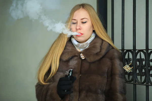 気まぐれな女だ毛皮のコートの若いかわいい女の子のブロンドは 電子タバコを吸い 冬に屋外でヴィンテージゲートウェイの近くに蒸気のパフをオフにすることができます — ストック写真