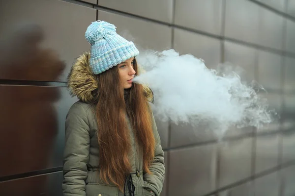 Vape genç. Genç güzel beyaz kız kış aylarında modern kahverengi zemin caddenin karşısında bir elektronik sigara içiyorum. Kötü alışkanlık. Yakın çekim.