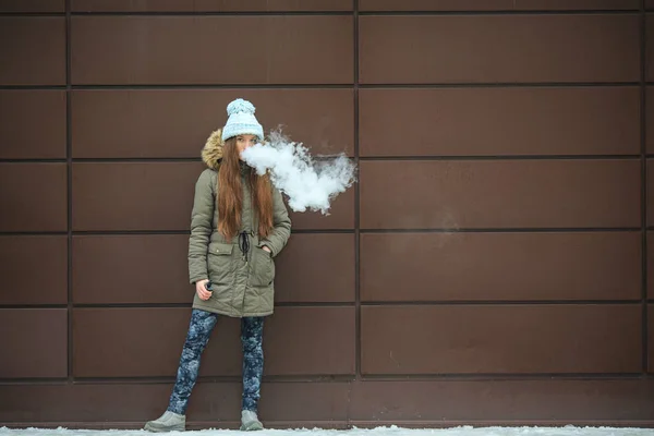 Vape genç. Genç güzel beyaz kız kış aylarında modern kahverengi zemin caddenin karşısında bir elektronik sigara içiyorum. Kötü alışkanlık.