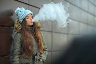Vape genç. Genç güzel beyaz kız mavi şapkalı kışın modern kahverengi zemin caddenin karşısında elektronik sigara sigara içiyor. Kötü alışkanlık. Yakın çekim.