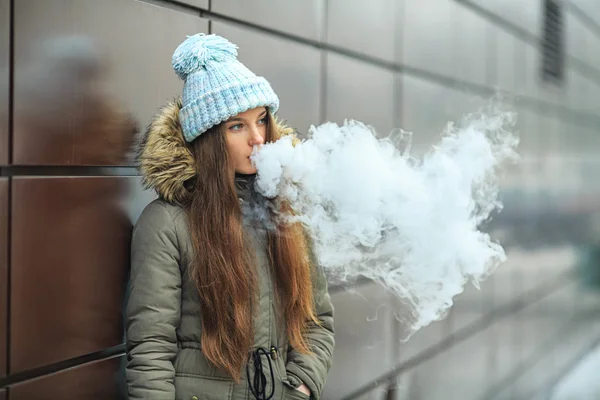 Vape genç. Genç güzel beyaz kız mavi şapkalı kışın modern kahverengi zemin caddenin karşısında elektronik sigara sigara içiyor. Kötü alışkanlık. Yakın çekim.