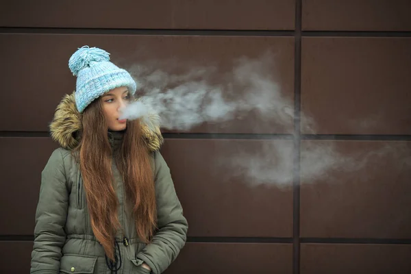 Vape genç. Genç güzel beyaz kız mavi şapkalı kışın modern kahverengi zemin caddenin karşısında elektronik sigara sigara içiyor. Kötü alışkanlık. Yakın çekim.