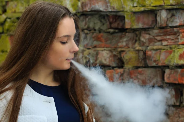 Vape genç. Genç güzel beyaz hırka baharda yıkılan tuğla duvar sokak karşısında elektronik sigara sigara içinde beyaz kız. Kötü alışkanlık.