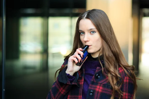 Vape genç. Genç güzel beyaz kız ters bir elektronik sigara kareli ceket cam sokakta baharda bina. Kötü alışkanlık. Vaping aktivite.