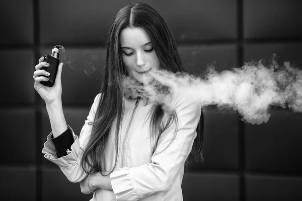 Vape genç. Gündelik giyim genç güzel beyaz kız bahar sokakta Modern arka plan karşısında bir elektronik sigara sigara. Kötü alışkanlık. Vaping aktivitesi. Kapat şunu. Siyah beyaz.