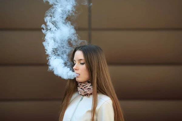 Vape teenager. Young pretty white girl in casual clothing smoking an electronic cigarette opposite modern brown background on the street in the spring. Bad habit. Vaping activity. Close up.