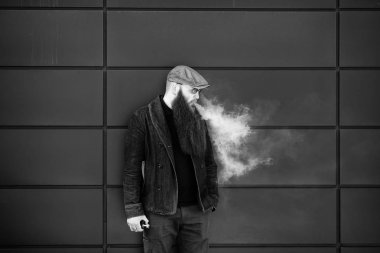 Vape sakallı adam. Bir şapka ve güneş gözlüğü çok uzun sakallı yetişkin bir adam güneşli bir gün dışında elektronik sigara içiyor. Sağlık için zararlı kötü alışkanlık. Siyah beyaz.