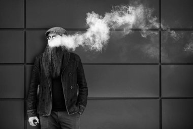 Vape sakallı adam. Bir şapka ve güneş gözlüğü çok uzun sakallı yetişkin bir adam güneşli bir gün dışında elektronik sigara içiyor. Sağlık için zararlı kötü alışkanlık. Siyah beyaz.