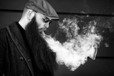 Vape sakallı adam. Bir şapka ve güneş gözlüğü çok uzun sakallı yetişkin bir adam güneşli bir gün dışında elektronik sigara içiyor. Sağlık için zararlı kötü alışkanlık. Siyah beyaz.