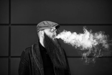 Vape sakallı adam. Bir şapka ve güneş gözlüğü çok uzun sakallı yetişkin bir adam güneşli bir gün dışında elektronik sigara içiyor. Sağlık için zararlı kötü alışkanlık. Siyah beyaz.