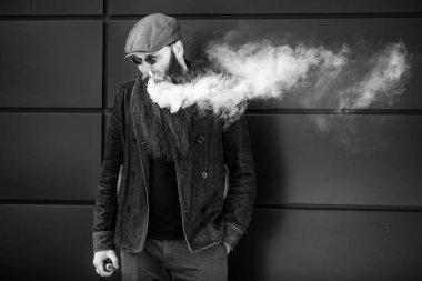 Vape sakallı adam. Bir şapka ve güneş gözlüğü çok uzun sakallı yetişkin bir adam güneşli bir gün dışında elektronik sigara içiyor. Sağlık için zararlı kötü alışkanlık. Siyah beyaz.