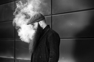 Vape sakallı adam. Bir şapka ve güneş gözlüğü çok uzun sakallı yetişkin bir adam güneşli bir gün dışında elektronik sigara içiyor. Sağlık için zararlı kötü alışkanlık. Siyah beyaz.