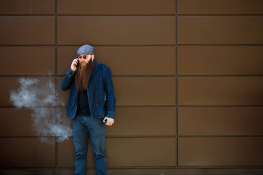 Vape sakallı adam. Bir şapka ve güneş gözlüğü çok uzun sakallı bir yetişkin adam dışında güneşli bir günde mobie telefonda elektronik bir sigara ve talkes sigara. Sağlığa zararlı kötü alışkanlık.