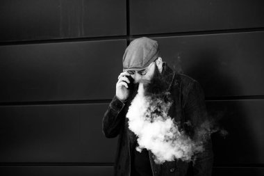 Vape sakallı adam. Bir şapka ve güneş gözlüğü çok uzun sakallı bir yetişkin adam dışında güneşli bir günde mobie telefonda elektronik bir sigara ve talkes sigara. Kötü alışkanlık. Siyah beyaz.