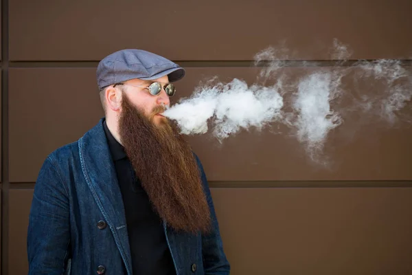 Vape sakallı adam. Bir şapka ve güneş gözlüğü çok uzun sakallı yetişkin bir adam güneşli bir gün dışında elektronik sigara içiyor. Sağlığa zararlı kötü alışkanlık.