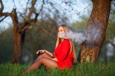 Vape kadını. Kırmızı elbiseli genç güzel sarışın kız bir ağacın yanında oturur ve baharda bir parkta açık havada gün batımında elektronik bir sigara içiyor. Sağlık için zararlı kötü alışkanlık. Vaping etkinliği.