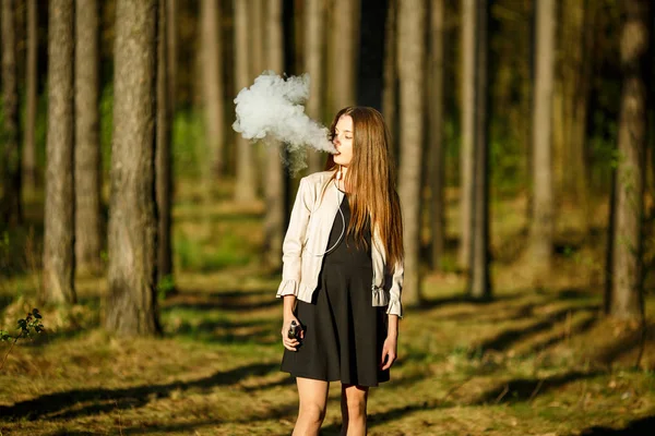 Vape teenager. Young cute girl in casual clothes smokes an electronic cigarette outdoors in the forest at sunset in summer. Bad habit that is harmful to health. Vaping activity.