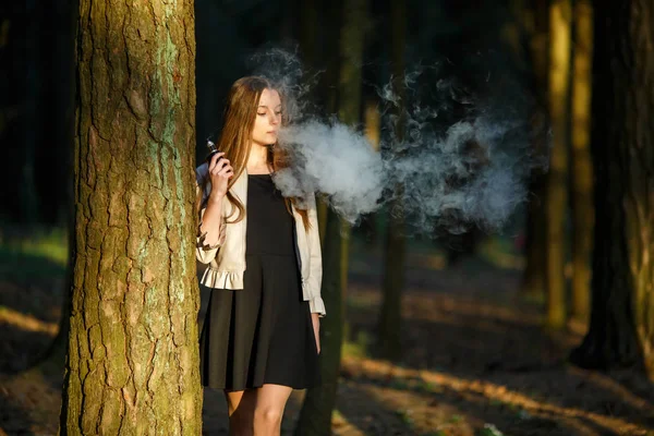 Vape genç. Rahat giysiler içinde Genç sevimli kız yaz aylarında gün batımında ormanda bir elektronik sigara açık sigara içiyor. Sağlık için zararlı kötü alışkanlık. Vaping etkinliği.