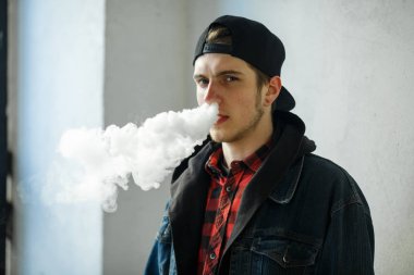 Vape genç. İlkbaharda sokakta elektronik sigara içen bir şapkalı genç yakışıklı adamın portresi. Sağlık için zararlı kötü alışkanlık. Vaping aktivitesi. Yakın çekim.