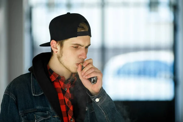 Vape genç. İlkbaharda sokakta elektronik sigara içen bir şapkalı genç yakışıklı adamın portresi. Sağlık için zararlı kötü alışkanlık. Vaping aktivitesi. Yakın çekim.