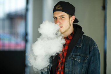 Vape genç. İlkbaharda sokakta elektronik sigara içen bir şapkalı genç yakışıklı adamın portresi. Sağlık için zararlı kötü alışkanlık. Vaping aktivitesi. Yakın çekim.