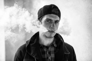 Vape genç. İlkbaharda sokakta elektronik sigara içen bir şapkalı genç yakışıklı adamın portresi. Sağlık için zararlı kötü alışkanlık. Vaping aktivitesi. Siyah ve beyaz. Yakın çekim.