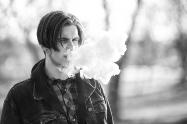 Vape genç. Genç yakışıklı adamın ilkbaharda bir parkta elektronik sigara içerken portresi. Sağlık için zararlı kötü alışkanlık. Vaping aktivitesi. Siyah beyaz.