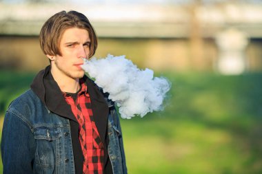 Vape genç. Genç yakışıklı adamın ilkbaharda bir parkta elektronik sigara içerken portresi. Sağlık için zararlı kötü alışkanlık. Vaping etkinliği.