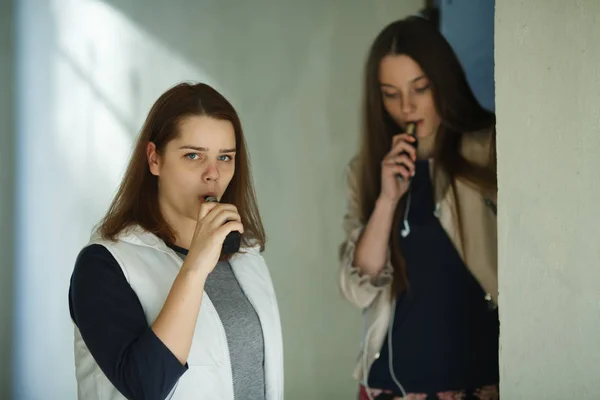 Vape teenagers. Two young cute girls in casual clothes smoke electronic cigarettes outdoors in the street in summer day. Bad habit that is harmful to health. Vaping activity.