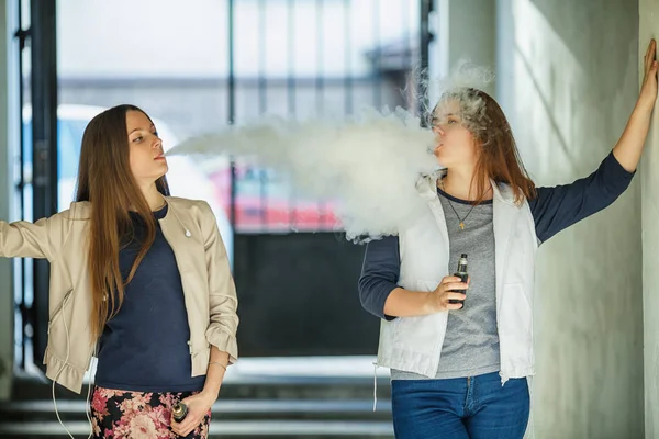 Vape gençleri. Gündelik giysiler içinde iki genç sevimli kız yaz günü sokakta elektronik sigara duman. Sağlık için zararlı kötü alışkanlık. Vaping etkinliği.
