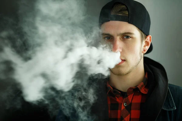 Vape genç. İlkbaharda sokakta elektronik sigara içen bir şapkalı genç yakışıklı adamın portresi. Sağlık için zararlı kötü alışkanlık. Vaping aktivitesi. Yakın çekim.