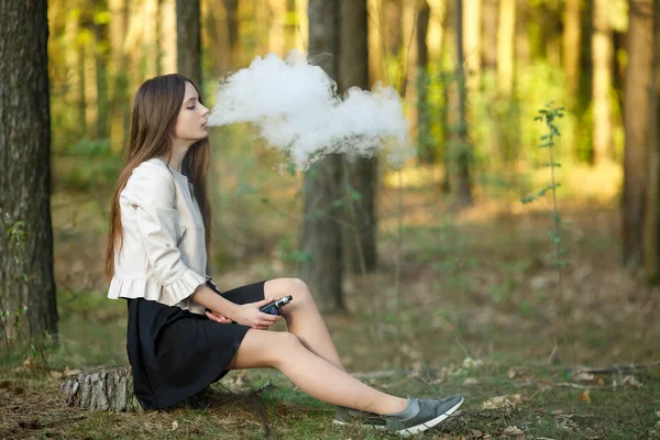 Vape teenager. Young cute girl in casual clothes smokes an electronic cigarette outdoors in the forest at sunset in summer. Bad habit that is harmful to health. Vaping activity.