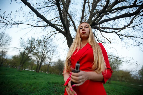 気まぐれな女だ赤いドレスを着た若い美しいブロンドの女の子は 春の公園で夜の屋外で大きな木の向かいに電子タバコを吸います 健康に有害な悪い習慣 — ストック写真