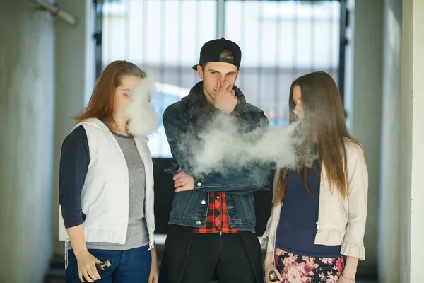 Vape gençleri. Gündelik giysiler içinde iki genç sevimli kız yaz günü sokakta sigara içmeyen adam yakınında elektronik sigara duman. Sağlık için zararlı kötü alışkanlık. Vaping etkinliği.