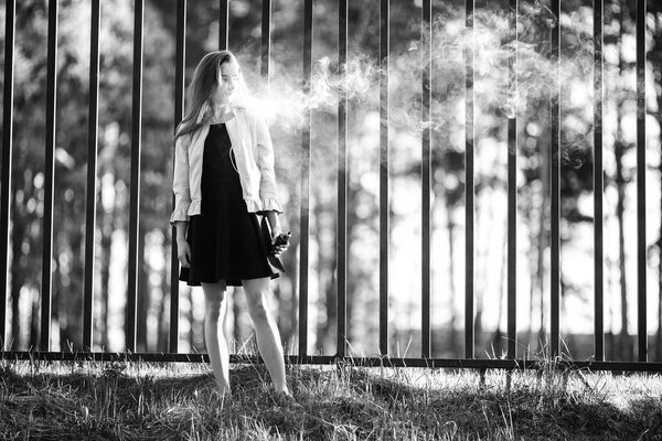 Vape teenager. Young cute girl in casual clothes smokes an electronic cigarette in front of a metal fence outdoors in the forest at sunset in summer. Bad habit. Stop vaping. Black and white.