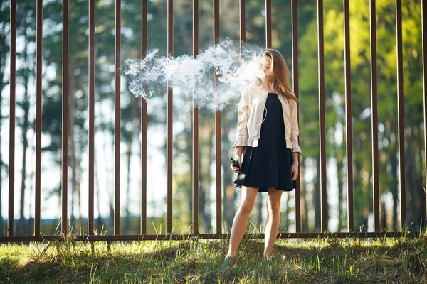Vape teenager. Young cute girl in casual clothes smokes an electronic cigarette in front of a metal fence outdoors in the forest at sunset in summer. Bad habit. Stop vaping.