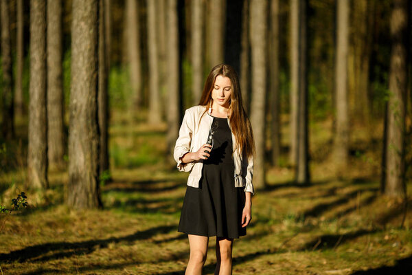 Vape teenager. Young cute girl in casual clothes smokes an electronic cigarette outdoors in the forest at sunset in summer. Bad habit that is harmful to health. Vaping activity.